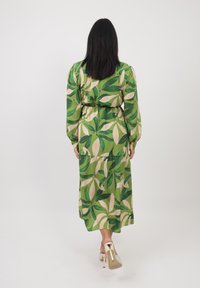 Abito verde con grandi motivi a foglia e petali crema; maniche lunghe, vita cinchata, tessuto leggero, lunghezza ankle, abbinato a scarpe con tacco chiaro.