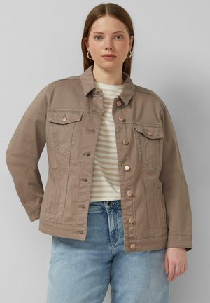 Młoda kobieta w mauve'owej zapinanej na guziki kurtce, pod którą ma beżową koszulę w paski, oraz w niebieskich dżinsach, stoi na tle jasnoszarego tła.