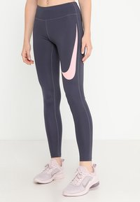 Leggings de Nike, gris oscuro con el swoosh rosa. Material elástico, diseño ajustado, costuras planas. Llevados con zapatillas deportivas de color rosa claro.