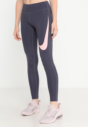 Leggings de Nike, gris oscuro con el swoosh rosa. Material elástico, diseño ajustado, costuras planas. Llevados con zapatillas deportivas de color rosa claro.