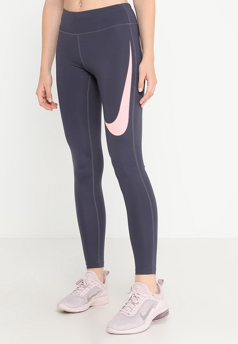Leggings de Nike, gris oscuro con el swoosh rosa. Material elástico, diseño ajustado, costuras planas. Llevados con zapatillas deportivas de color rosa claro.