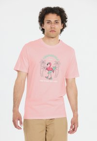 Vaaleanpunainen puuvillapaita, jossa on graafinen design flamingosta surffaamassa laudalla, ympärillä palmuja ja tekstiä kontrastiväreissä.