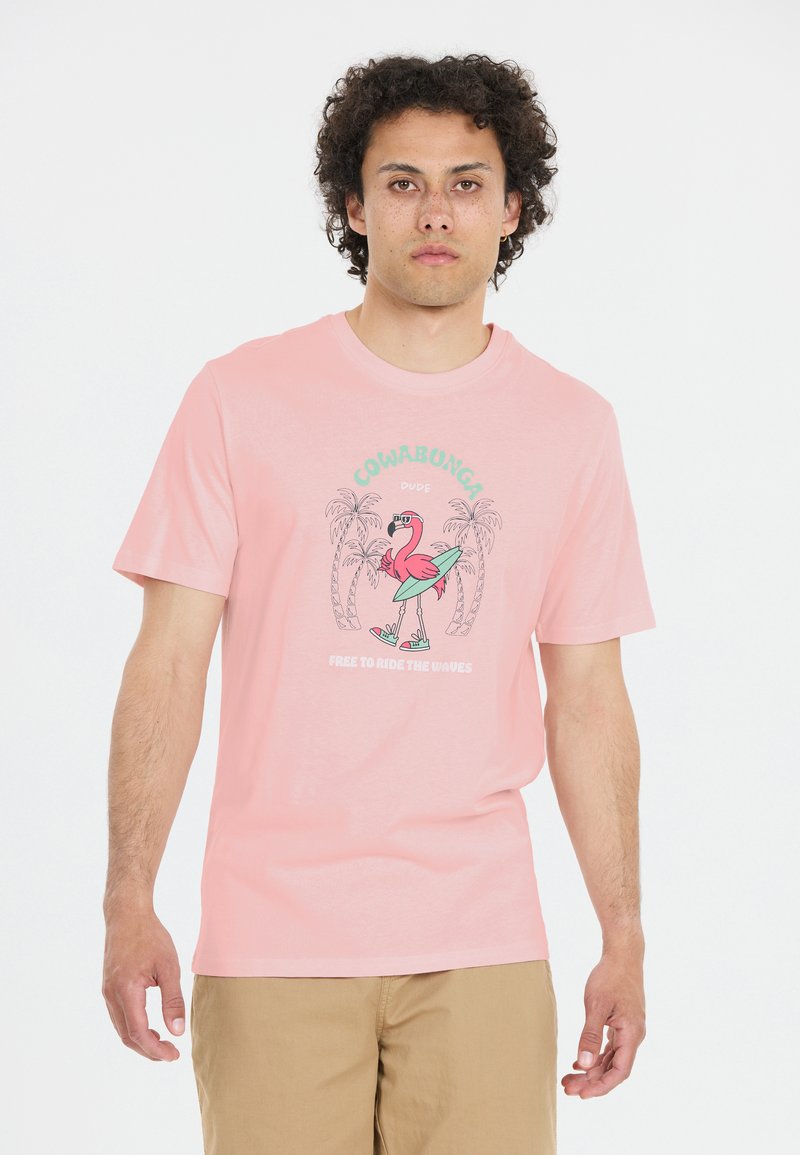 Vaaleanpunainen puuvillapaita, jossa on graafinen design flamingosta surffaamassa laudalla, ympärillä palmuja ja tekstiä kontrastiväreissä.