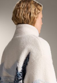 Persoană cu păr blond scurt și ondulat, văzută din profil, purtând o jachetă sherpa albă cu modele albastre în formă de stea pe spate.