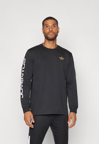 Camiseta negra de manga larga con un logo dorado en el pecho y texto blanco en la manga. Fabricada con un tejido suave y con un corte relajado.