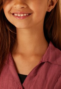 Goldene Marienkäfer Ohrringe mit roten Akzenten, getragen von einem Kind. Das Kind hat lange braune Haare und trägt ein strukturiertes mauvefarbenes Shirt.