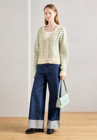 Cardigã de malha com um padrão de onda em menta e creme, apresentando botões de pérola. Combinado com jeans de perna larga e uma mala azul-clara.