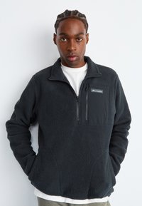 Zwarte fleece trui met een ritszak op de borst en een logo. Heeft een hoge kraag en een ontspannen pasvorm. Gestructureerde, zachte stof.