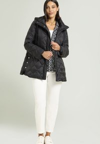 Donna con un giubbotto trapuntato nero sopra una maglia a fantasia, pantaloni bianchi e sneakers bianche, in piedi contro uno sfondo chiaro e semplice.