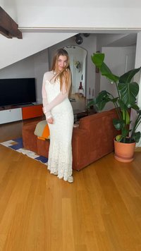 Mladá žena v bielych čipkových šatách stojí s úsmevom v moderne zariadenej obývačke s drevenou podlahou, hnedou pohovkou a veľkou kvetináčovou rastlinou.