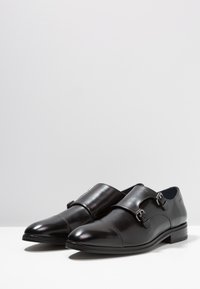 Zwarte leren dubbele monk strap schoenen met een spitse neus, een gladde afwerking, gespen aan elke kant en een lichte hak.