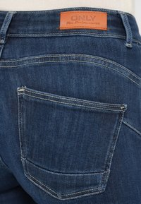 Κοντινή λήψη πίσω τσέπης μπλε τζιν με αντίθετη ραφή και καφέ δερμάτινη ετικέτα που γράφει "ONLY Blue Denim anno 1995."