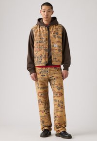 Chaleco y pantalones de color tostado con un estampado vintage, que presentan gráficos y texto coloridos. El chaleco está superpuesto a una sudadera oscura y una camisa roja.