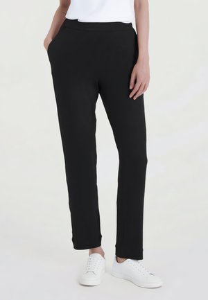 Persona indossa pantaloni neri a gamba dritta con tasche e scarpe da ginnastica bianche, in piedi davanti a uno sfondo chiaro e uniforme.