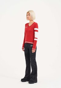 Roter V-Ausschnitt-Pullover mit weißen Streifen an den Ärmeln, kombiniert mit schwarzen Schlaghosen mit strukturiertem Muster und schwarzen Plateauschuhen.