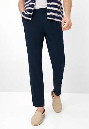 Mand iført marineblå afslappede bukser, beige slip-on sko, en hvid t-shirt og en mønstret navyblå og hvid vest, stående mod en hvid baggrund.