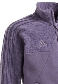 Lila fleece-jacka med hög krage, med texturerad tyg, släta detaljer och tre horisontella ränder längs ärmarna.