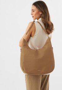 Borsa a spalla in pelle marrone chiaro con forma arrotondata, superficie testurizzata e dettagli in metallo dorato, che presenta un logo del marchio ben visibile.