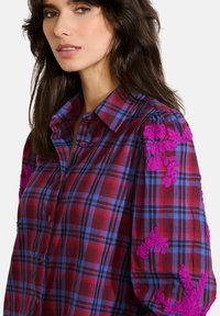 Červeno-modrá kostičkovaná košile s knoflíkovým zapínáním. Obsahuje živé purpurové květinové výšivky na rukávech a jemnou texturu.