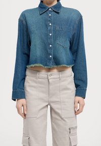 Camicia di jeans tagliata con bottone frontale, colletto, tasca sul petto e orlo sfilacciato in un lavaggio blu medio. Abbinata a pantaloni cargo di colore chiaro.