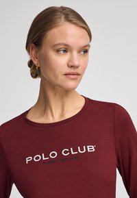 Marin långärmad topp med "POLO CLUB" broderat i vitt och blått, med rund halsringning och mjuk bomullstextur.