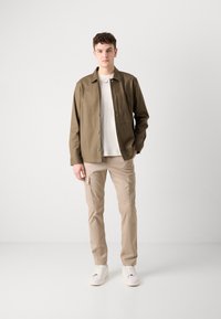 Brun knappad skjorta över en vit T-shirt, i kombination med beige cargo-byxor och vita sneakers, vilket skapar en avslappnad och funktionell outfit.