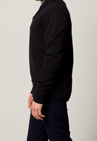 Svart stickad tröja med ribbad nederkant och ärmar, med en sjalkrage. Bärs över marinblå byxor, vilket visar en sidoprofil.