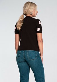 Kobieta z blond kucykiem, ubrana w czarny sweter z krótkim rękawem i różowymi kwiatowymi wzorami na rękawach oraz niebieskie dżinsy, zwrócona plecami.