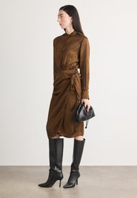 Abito marrone a fantasia con maniche lunghe e design avvolgente, abbinato a stivali neri con tacco al ginocchio e una piccola borsa nera.