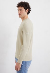 Helles beiges, geripptes Sweatshirt mit langen Ärmeln, Rundhalsausschnitt und strukturiertem Stoff. Verfügt über ein kleines Logo-Patch am linken Ärmel.