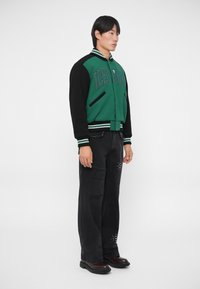 Giacca varsity verde e nera con maniche a contrasto, colletto rigato, lettere ricamate, abbinata a jeans neri strappati e scarpe nere.