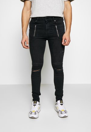 Jeans neri aderenti con tasche frontali con cerniera, ginocchia strappate e scarpe da ginnastica bianche con accenti gialli indossate da una persona in piedi su uno sfondo neutro.