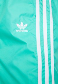 Tyrkysová bunda Adidas se bílým designem tří pruhů a vyšívaným logem na levé straně, vyrobená z hladkého, lehkého materiálu.