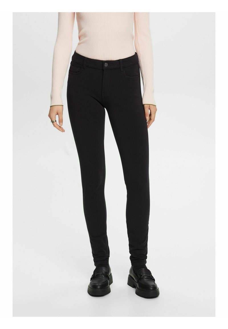 Femme portant un pantalon skinny noir, des mocassins noirs et un haut côtelé à manches longues rose clair, debout devant un fond uni.