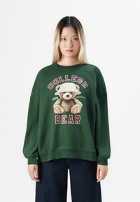 Giovane donna che indossa un felpa verde scuro con un orsetto in cappello a secchiello e le parole "College Bear" in rosso e bianco.