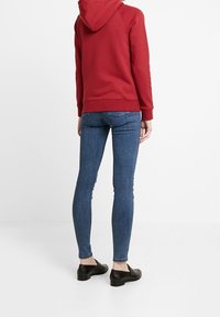 Sudadera roja con capucha, combinada con jeans ajustados azules. Zapatos negros resaltan el conjunto casual. Enfócate en los materiales y el ajuste.