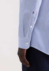 Camicia a righe blu e bianche con polsino abbottonato, caratterizzata da un piccolo dettaglio ricamato sulla manica e da una texture tessile liscia.