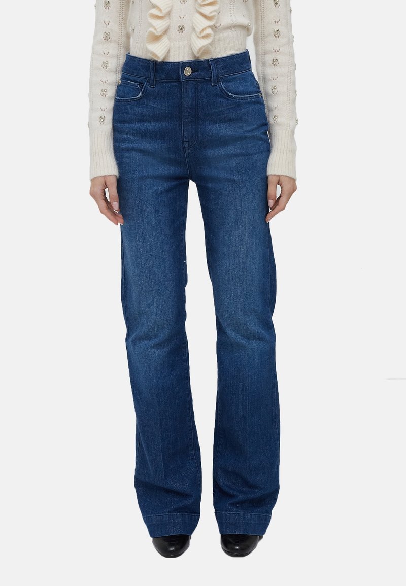 Femme portant un jean bleu taille haute à jambes droites, associé à un pull en maille crème orné de volants et de détails en perles.