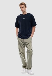 Camiseta de algodón azul marino de mangas cortas, con un pequeño parche de logo; combinada con pantalones cargo beiges claros con bolsillos laterales y deportivas blancas.