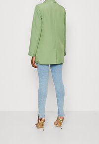 Blazer lungo verde con taglio sartoriale, abbinato a jeans skinny azzurri e sandali con tacco alto e motivo floreale.