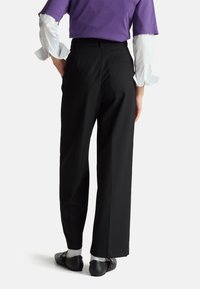 Zwarte brede broek met een gladde textuur, voorzien van achterzakken. Gecombineerd met zwarte platte schoenen en zichtbare witte sokken.
