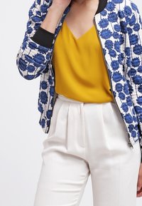 Vadderad bomberjacka i vitt med blå blommönster, tillsammans med en senapsgul V-ringad topp och vita plisserade byxor.