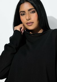 Frau mit langem schwarzem Haar, die einen schwarzen Pullover trägt und den Kragen des Pullovers vorsichtig nahe an ihr Gesicht hält, vor einem weißen Hintergrund.