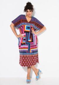 Bunt gemustertes Kleid mit geometrischem Muster in Pink, Blau und Schwarz, mit V-Ausschnitt und kurzen Ärmeln. Kombiniert mit blauen Blockabsatzschuhen.
