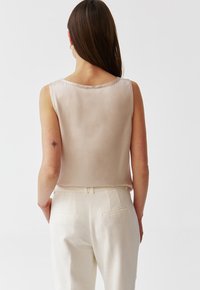 Beżowy bezrękawnik z satyny z okrągłym dekoltem, gładką teksturą i subtelnym połyskiem, zestawiony z jasnymi, dopasowanymi spodniami.