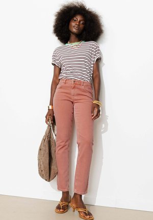Femme aux cheveux afro naturels portant un t-shirt rayé, un jean rose taille haute, des sandales jaunes, des bracelets en or et tenant un sac marron.