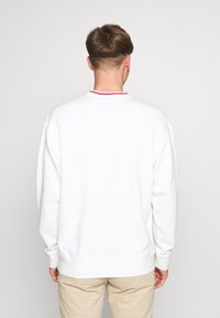 Sudadera blanca con cuello redondo y acento rojo en el escote. Presenta hombros caídos y puños de canalé. Tejido suave y texturizado.