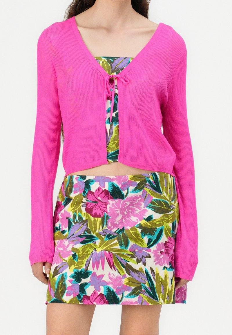 Femme portant un cardigan court rose vif noué à la poitrine, par-dessus un haut à motif floral et une mini-jupe assortie à motif floral.