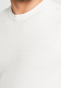 Vit kortärmad skjorta med en ribbad textur och rundad halsringning, som har subtila horisontella ränder genom hela tyget.