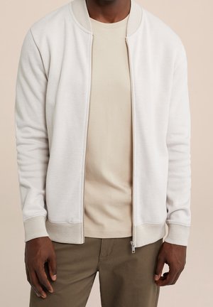 Mann trägt eine hellbeige, texturierte Jacke mit Reißverschluss über einem beigen T-Shirt und olivgrünen Hosen, steht vor einem einfarbigen Hintergrund.